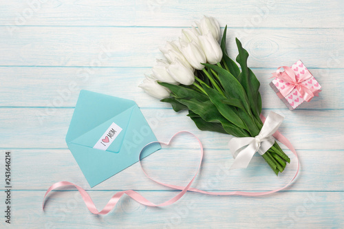 Fototapeta Naklejka Na Ścianę i Meble -  A bouquet of white tulips and a pink ribbon in the form of a heart with a gift box, love note and color envelope on blue wooden boards. Mother's day