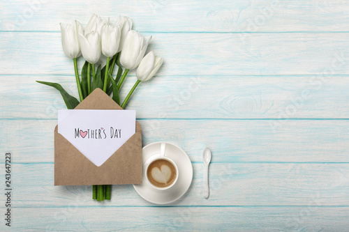 A bouquet of white tulips, cup of coffee with a love note and envelope on blue wooden boards . Mother's day