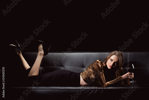 beautiful elegant woman lying on couch with glass of red wine while looking at camera isolated on black