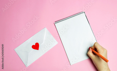 Female hand writing a love letter. Valentines day concept. Greeting valentine card. Declaration of love. Wedding invitation. Red heart. Pink background. View from above. Flat lay