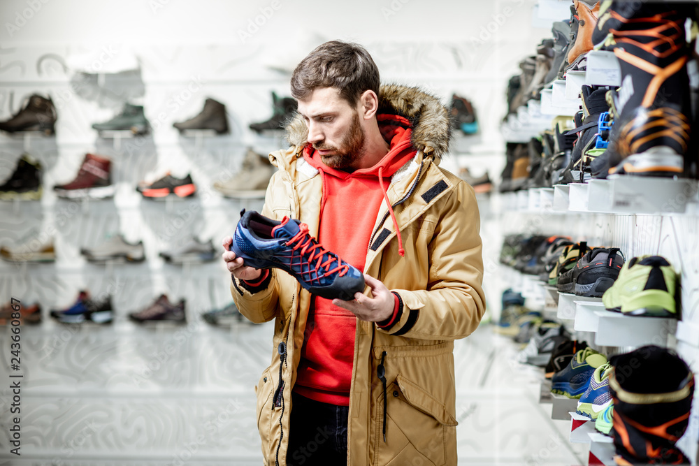 Man in winter jacket choosing trail shoes for mountain hiking in the sports shop
