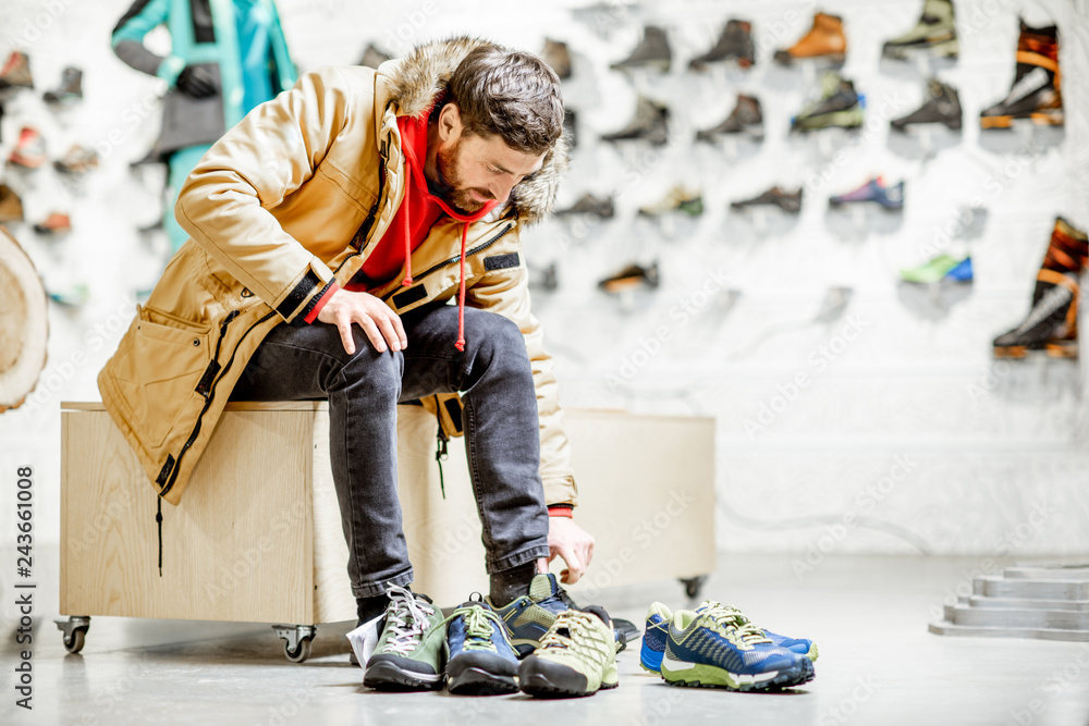 Man in winter jacket trying shoes for mountain hiking sitting in the fitting room of the modern sports shop - obrazy, fototapety, plakaty 