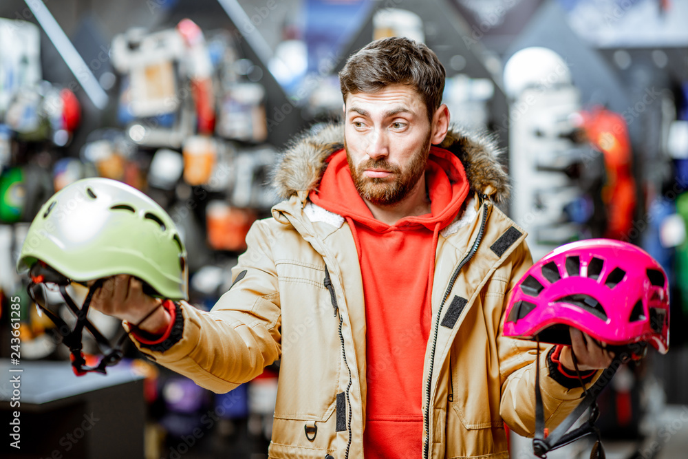 Man in winter jacket choosing mountaineer equipment holding helmets for hiking in the sports shop