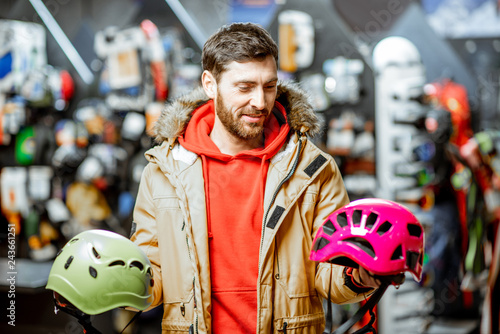 Man in winter jacket choosing mountaineer equipment holding helmets for hiking in the sports shop