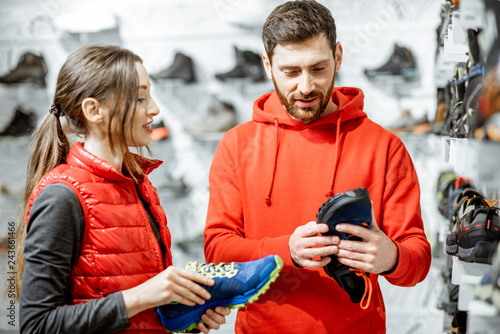 Mam and woman in red sports clothes choosing trail shoes for hiking standing near the showacase of the modern sports shop