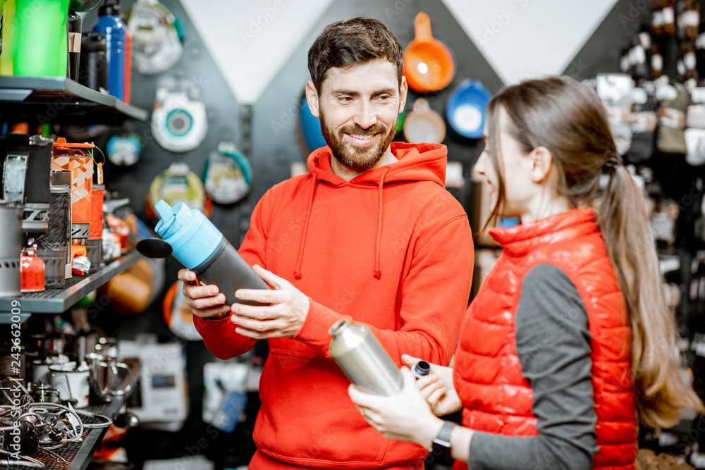 Young couple dressed in red sportswear choosing dishes for camping in the shop with travel equipment