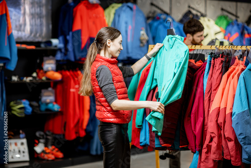 Young woman choosing winter clothes picking up down jackets on the hanger in the sports shop