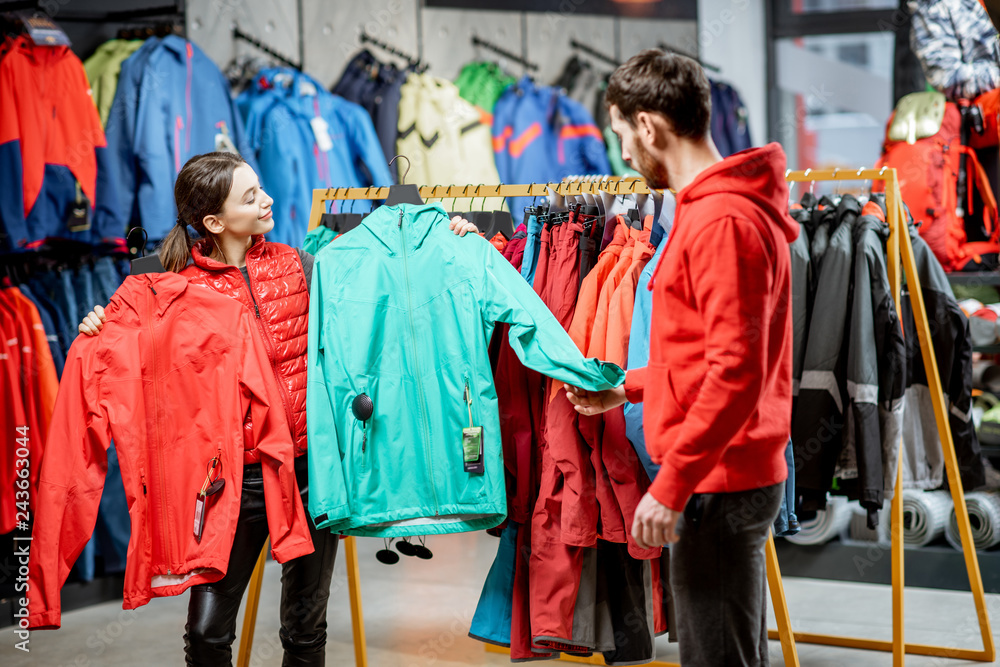 Young woman choosing between red and turquoise jacket standing with boyfriend in the sports shop