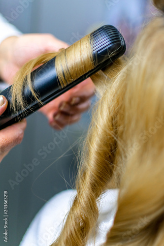 Close-up photo professional hairdresser uses flat iron for making curls on beautiful blonde hair