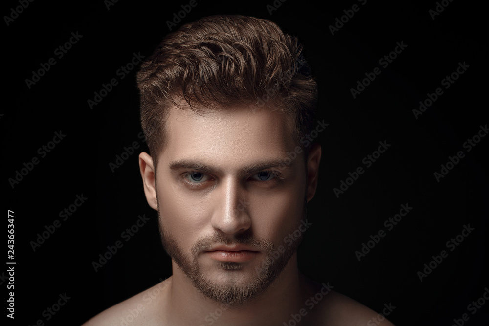 Closeup view portrait of one handsome bearded young macho man with strong look hazel eyes and sexy lips standing in light looking forward in studio on black background