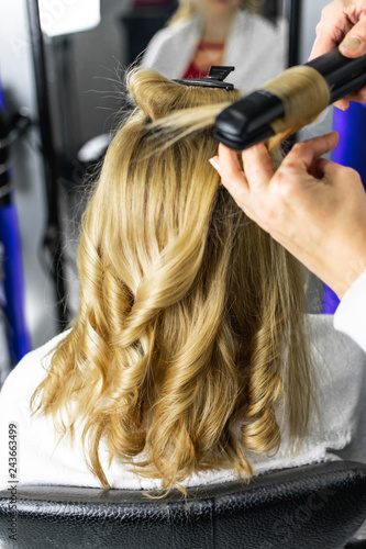 Close-up photo professional hairdresser uses flat iron for making curls on beautiful blonde hair. Client sitting on the chair