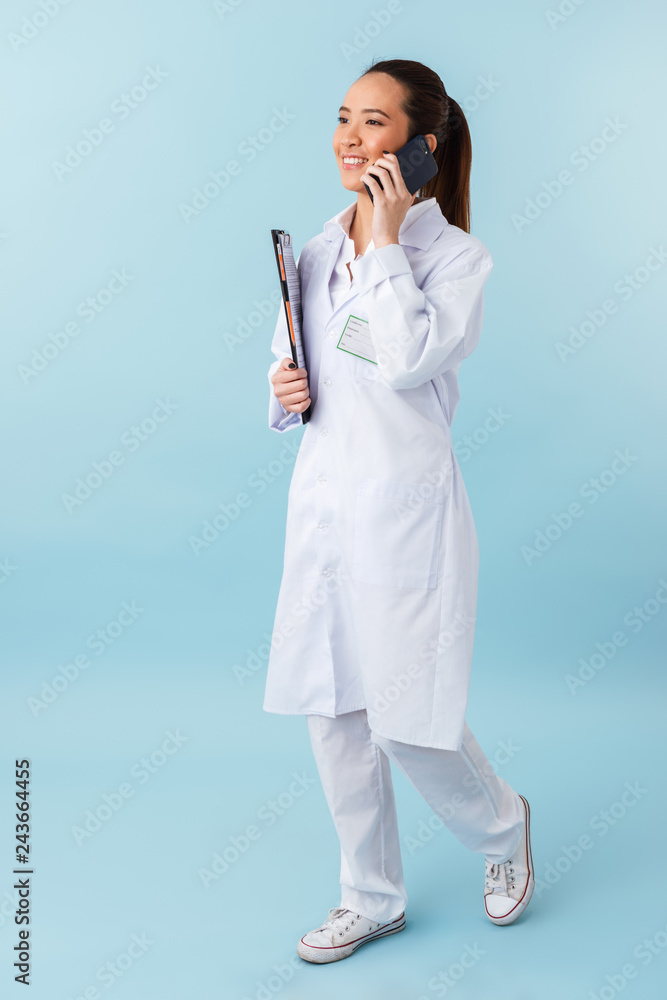 Woman doctor posing isolated over blue wall background holding clipboard talking by mobile phone. Woman doctor posing isolated over blue wall background holding clipboard talking by mobile phone.