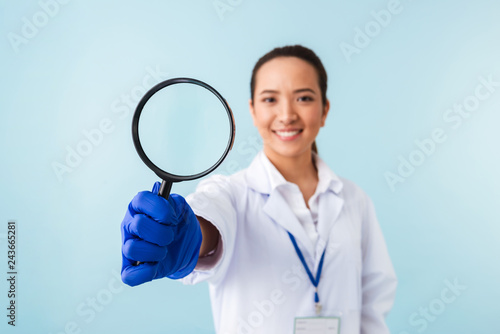 Woman doctor posing isolated over blue wall background holding magnifying glass.