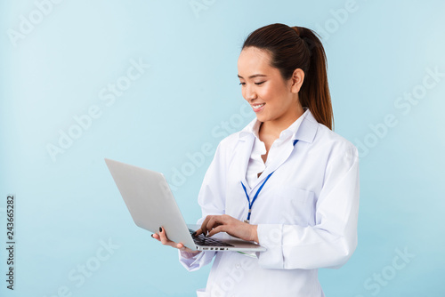 Young woman doctor posing isolated over blue wall background using laptop computer.