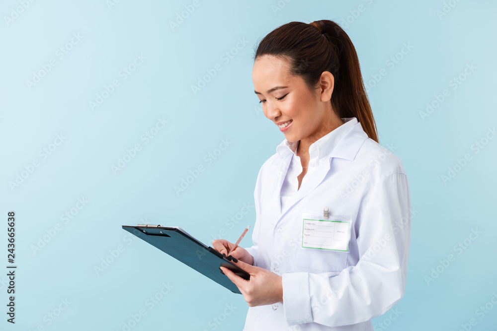 Cheerful young woman doctor posing isolated over blue wall background holding clipboard. Cheerful young woman doctor posing isolated over blue wall background holding clipboard.