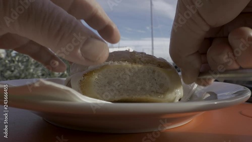 Man's hand picks up a knife and cuts into a cinnamon bun then places the knife back onto the plate at a harbour cafe on a sunny day with yachts in the background shown as a big close-up in slow motion