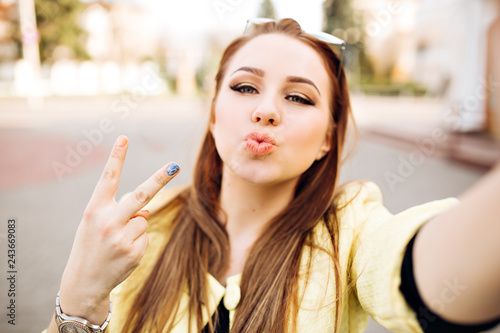 Young sexy woman with bright make-up formic blond hair makes selfie sending a kiss, hipster street portrait. Shows a peace sign, taking pictures of herself