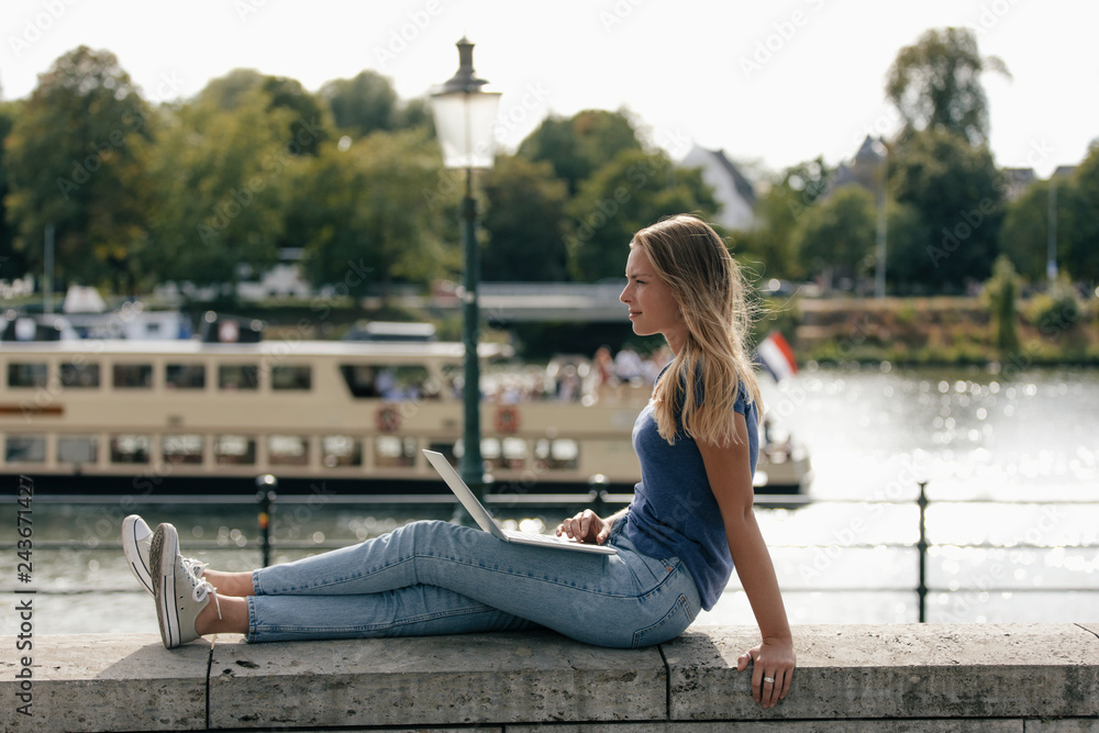 Netherlands, Maastricht, young woman sitting on a wall at the riverside with laptop Netherlands, Maastricht, young woman sitting on a wall at the riverside with laptop