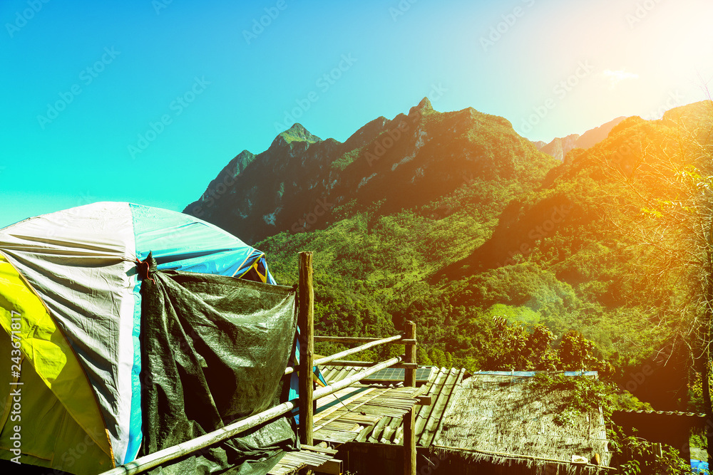 A large camping tent located in front of a large mountain with green trees covered. Do not be rich in the sunlight that is shining through and the blue sky.