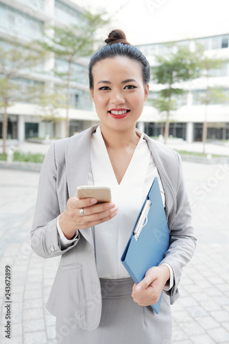 Portrait of cheerful attractive businesswoman with smartphone standing outdoors and smiling at camera Free Download Portrait of cheerful attractive businesswoman with smartphone standing outdoors and smiling at camera - GFXLoad