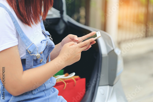 young woman hand holding smartphone and shopping bags with standing at the car parking lot, shopping concept