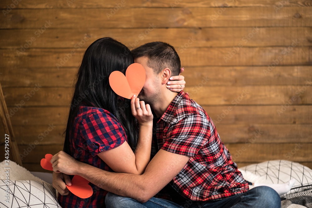 Romantic couple sitting on a sofa in a loving embrace kissing as they hold  up a red paper heart on wooden wall  kissing behind the  heart for Valentines day Stock Photo |