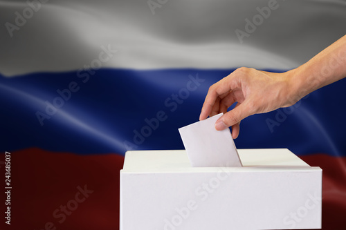 Close-up of human hand casting and inserting a vote and choosing and making a decision what he wants in polling box with Russia flag blended in background.