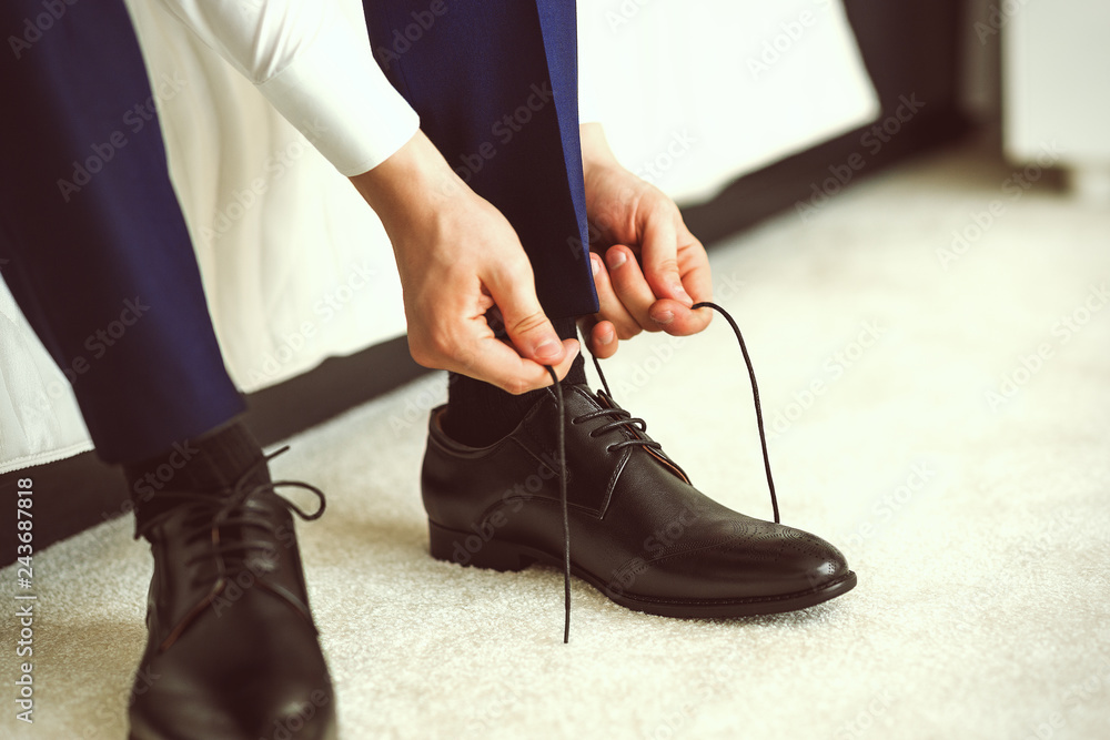 A man in a blue suit ties up shoelaces on black classic elegant shoes. Groom morning in hotel room before wedding ceremony.