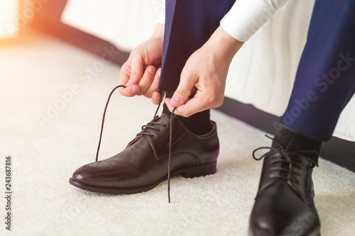 A man in a blue suit ties up shoelaces on black classic elegant shoes. Groom morning in hotel room before wedding ceremony.