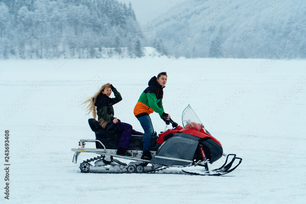 Man and woman riding on a snowmobile on the frozen lake in the mountains with the scenic view. Pine trees covered with snow. Side view