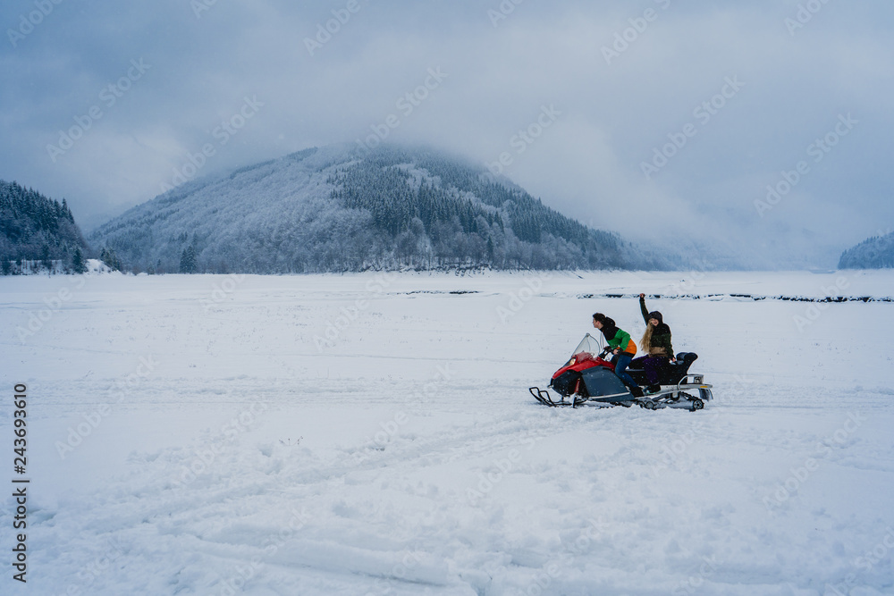 Man and woman riding on a snowmobile on the frozen lake in the mountains with the scenic view. Pine trees covered with snow. Side view