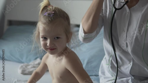 A doctor in the hospital is listening to a little girl with a stetus. The baby is crumpled, the woman in the white coat smoothes the baby on the back. Pediatrician examination at home