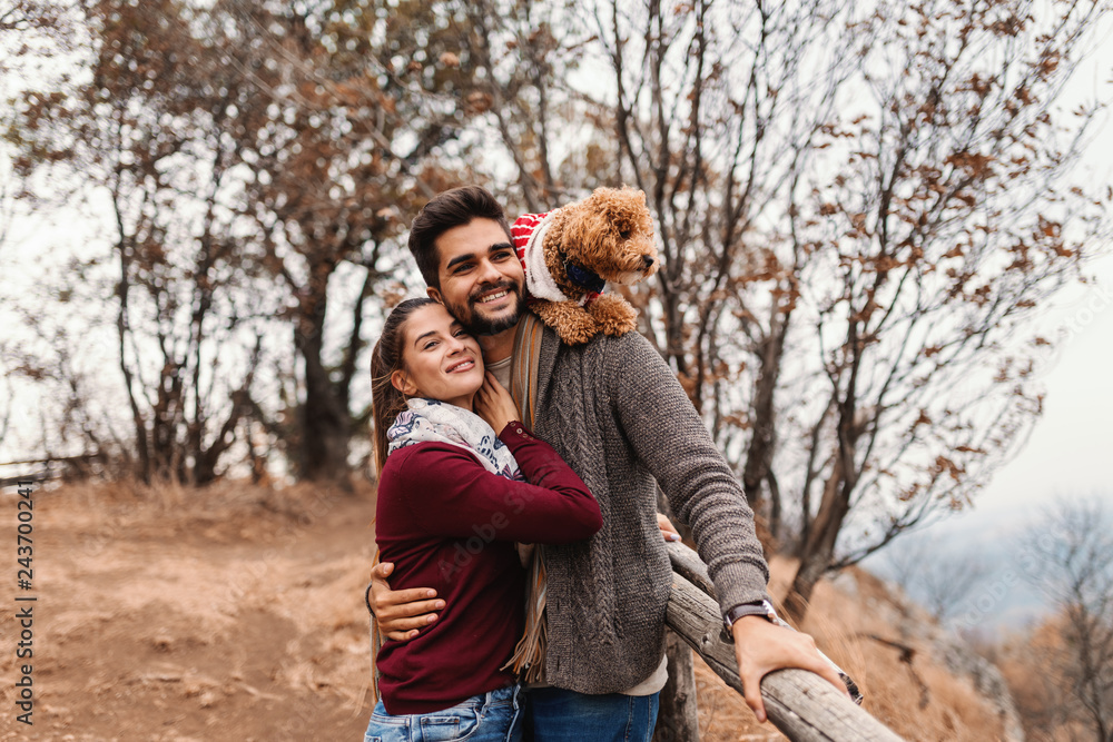Happy couple in love hugging and looking at beautiful view. Man holding on the back their apricot poodle. Autumn time. Happy couple in love hugging and looking at beautiful view. Man holding on the back their apricot poodle. Autumn time.