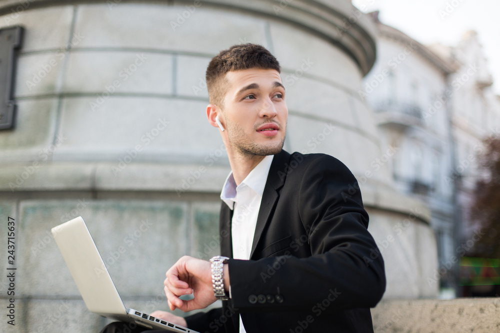 Young pensive businessman in black suit with wireless earphones dreamily looking aside with hand watch and laptop on knees outdoor Young pensive businessman in black suit with wireless earphones dreamily looking aside with hand watch and laptop on knees outdoor