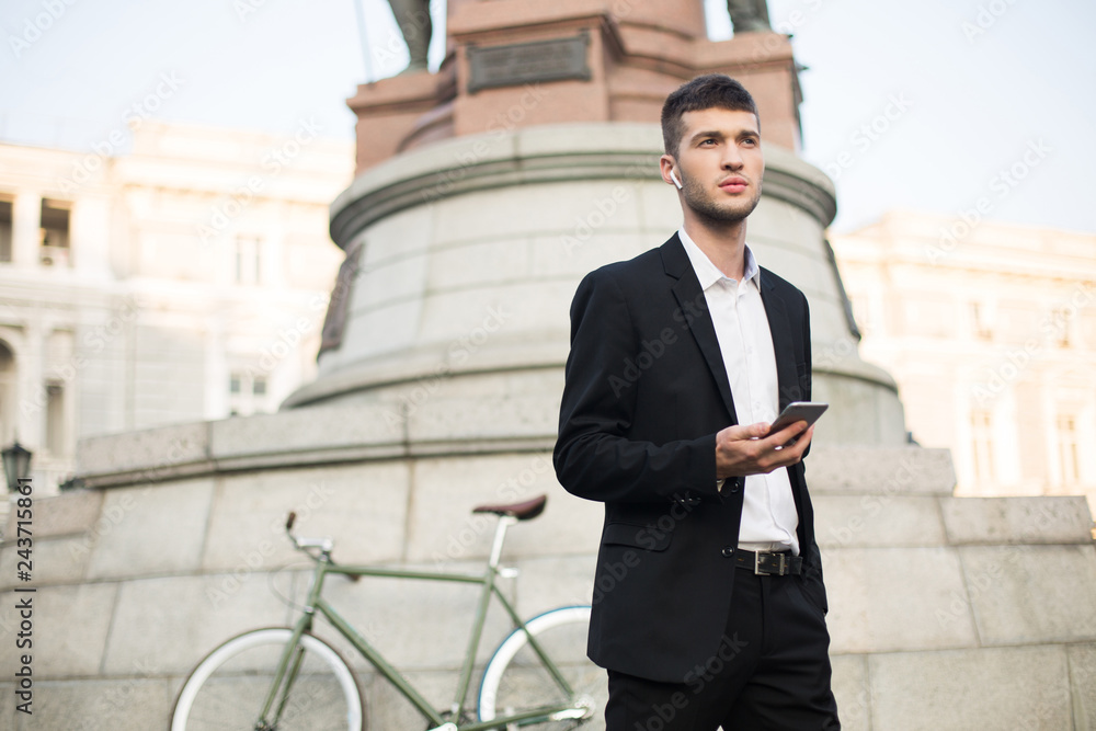 Young serious businessman in classic black suit and white shirt with wireless earphones holding cellphone in hand while thoughtfully looking aside with retro bicycle on fuzzy background outdoor Young serious businessman in classic black suit and white shirt with wireless earphones holding cellphone in hand while thoughtfully looking aside with retro bicycle on fuzzy background outdoor