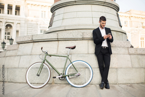 Young handsome man in classic black suit and white shirt with wireless earphones dreamily using cellphone with retro bicycle near on city street