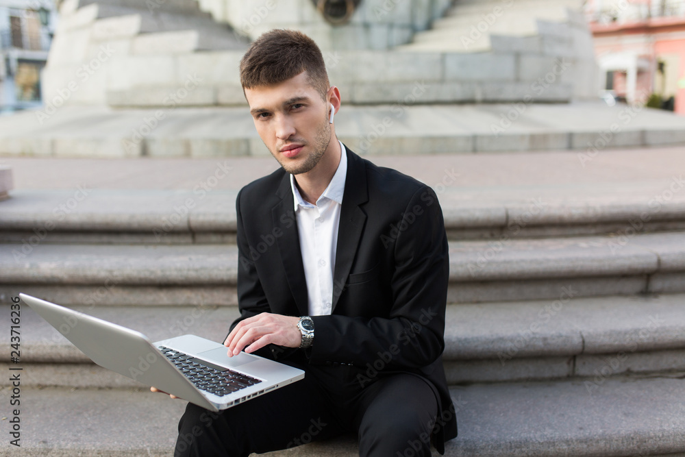 Young handsome businessman in classic black suit with wireless earphones thoughtfully looking in camera while holding laptop in hands outdoor Young handsome businessman in classic black suit with wireless earphones thoughtfully looking in camera while holding laptop in hands outdoor