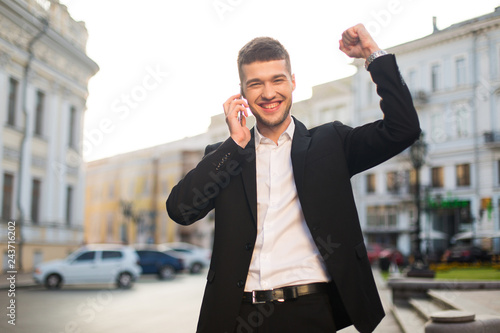 Young beautiful smiling man in black jacket and white shirt happily looking in camera raising hand up while talking on cellphone with city view on background