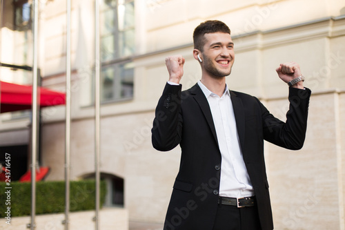 Young joyful businessman in black jacket and white shirt happily looking aside showing yes gesture while spending time outdoor
