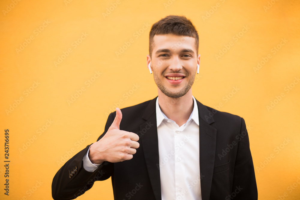 Young smiling man in classic black jacket and white shirt with wireless earphones happily showing thumb up gesture while looking in camera over orange background Young smiling man in classic black jacket and white shirt with wireless earphones happily showing thumb up gesture while looking in camera over orange background