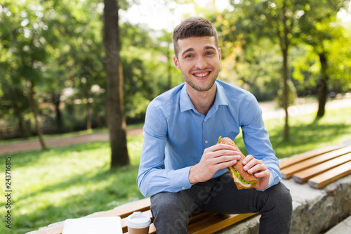 Young joyful man in blue shirt sitting on bench with sandwich in hands and cup of coffee to go near happily looking in camera while spending time in cozy park