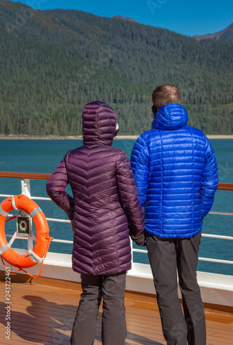 Couple in colorful purple and blue winter puffer jackets view Alaska, USA coastl from ship's deck on a sunny cold day. photo