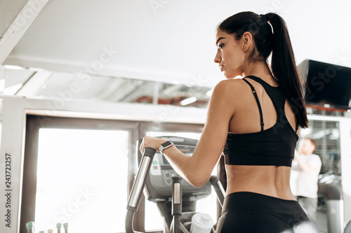 Dark-haired athletic girl dressed in black sports top and tights works out on a treadmill in the gym