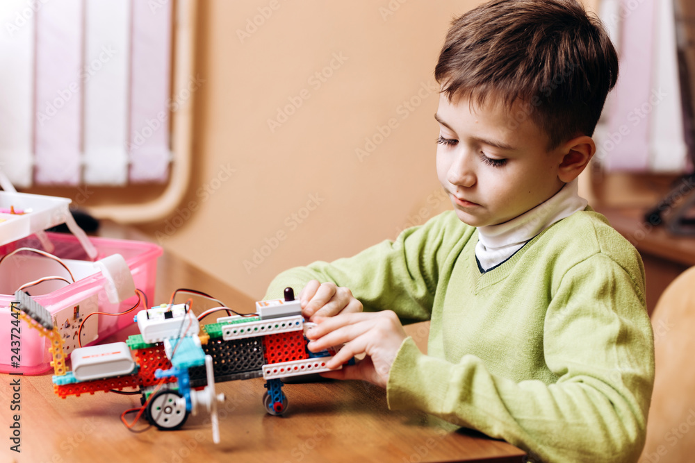 Diligent boy dressed in green sweater sits at the desk with computer and makes the robot from the robotic constructor in the School of Robotics Diligent boy dressed in green sweater sits at the desk with computer and makes the robot from the robotic constructor in the School of Robotics