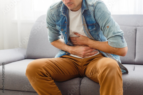 The man is sitting on a gray couch and holding his belly. Medicine and health concept, stomach problems. The man suffers from stomach ache, gastric problems. Abdominal pain, suffering and pain. photo