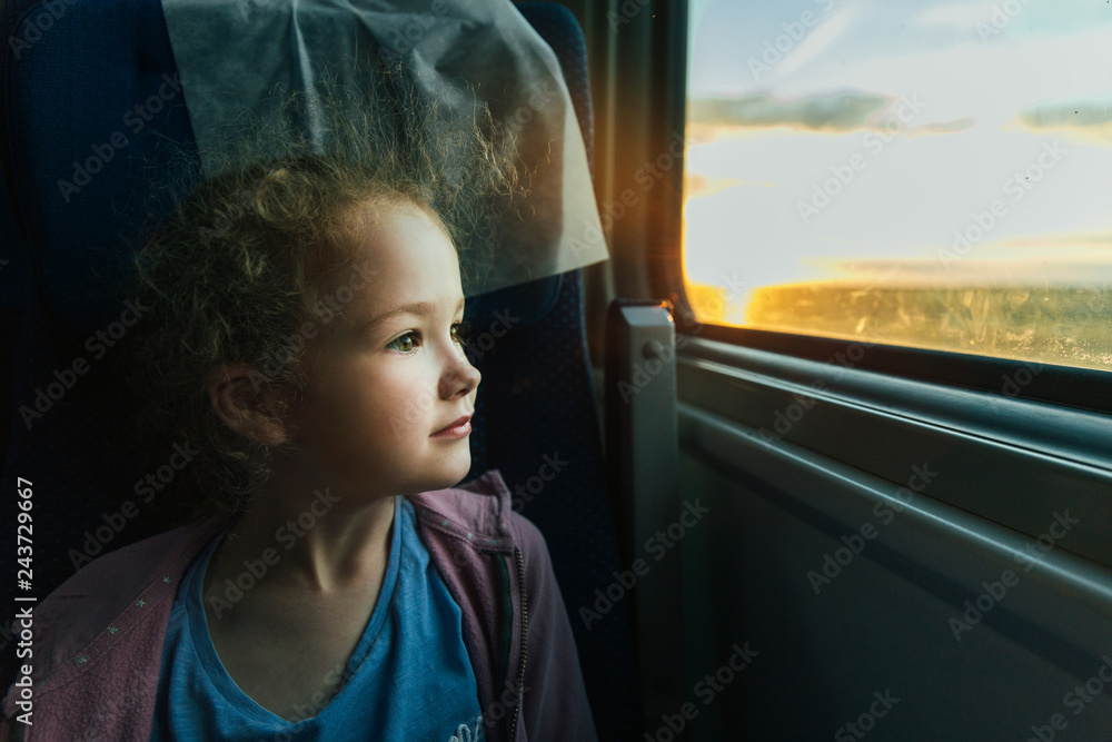 Beautiful little girl looking out train window outside, while it moving. Going on vacations and traveling by railway in summer. Sunset time.