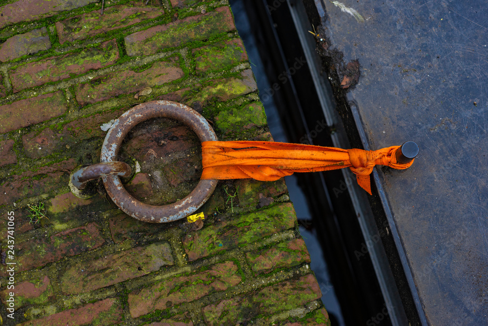 Fototapeta premium Rusty metal ring and boat tied with orange ribbon to the canal shore