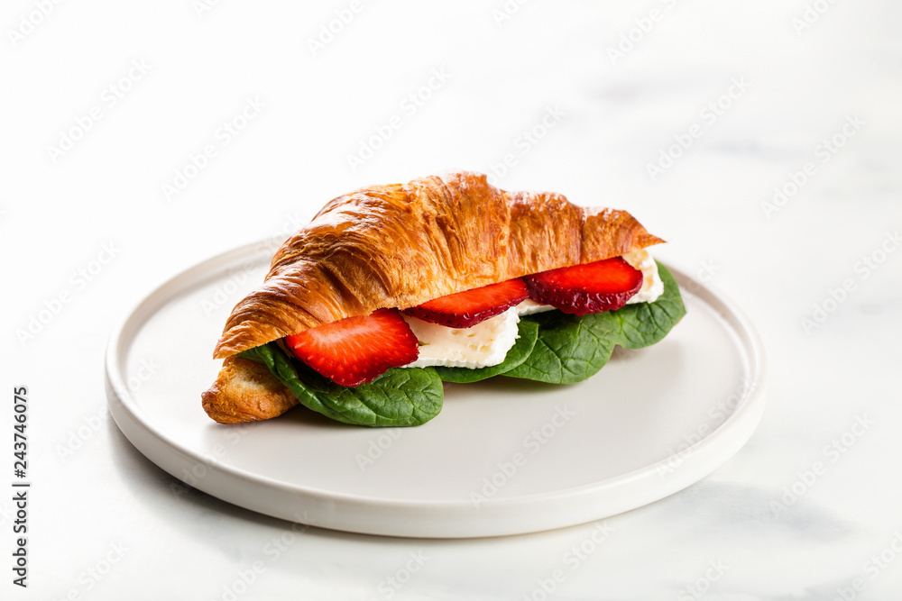 Fresh sandwich with croissant with cheese, strawberries, spinach on white plate.