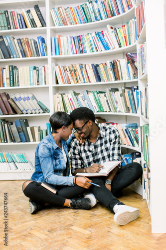 Happy young Afro-American couple sitting on a floor at the bookshelf in a library, smiling and reading book in library