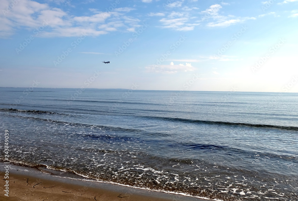 Seascape with passenger airplane flying in the sky with light clouds above calm sea and sandy beach on sunny day